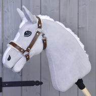 The Hobby Horse LarDen Halter with fur - leatherette Walnut is a gray plush hobby horse with a white mane, brown leatherette bridle, and large black eyes on a wooden stick, shown leaning against a gray wooden wall.