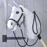 Western Teugels Montana van Hobby Horse LarDen, een grijze pluche hobby horse met witte manen en bles, zwart hoofdstel met musketonhaken en westernteugels, gemonteerd op een stevige houten stok en getoond op een grijze houten wand.