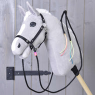 Gespleten Western Teugels Texas van Hobby Horse LarDen, een grijs pluche paard met witte manen, zwart hoofdstel, gespleten westernteugels en kleurrijke neklinten, gemonteerd op een houten stok voor een grijze houten deur.