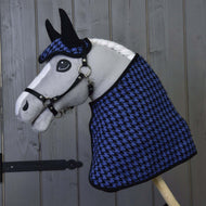 Een Hobby Horse LarDen hobby horse met witte manen en zwart hoofdstel draagt de Set feestelijk geruit blauw (oornetje en deken met pied-de-poulepatroon) en poseert voor een grijze houten wand.