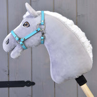 Halster - zwart kunstleer (maat S) van Hobby Horse LarDen op een grijze pluchen Hobby Horse met witte manen en blauw hoofdstel, gemonteerd op een houten stok voor grijze houten panelen.
