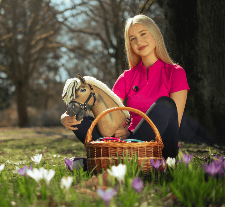 A young woman in a bright pink shirt sits on grass outdoors, holding a toy hobby horse and a wicker basket filled with colorful eggs, surrounded by blooming white and purple flowers.