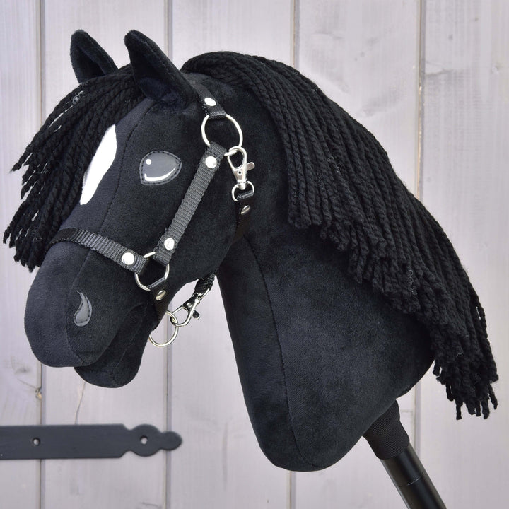 A black plush hobby horse with long yarn mane, white facial markings, and a black bridle, mounted on a stick, in front of a light wooden background.
