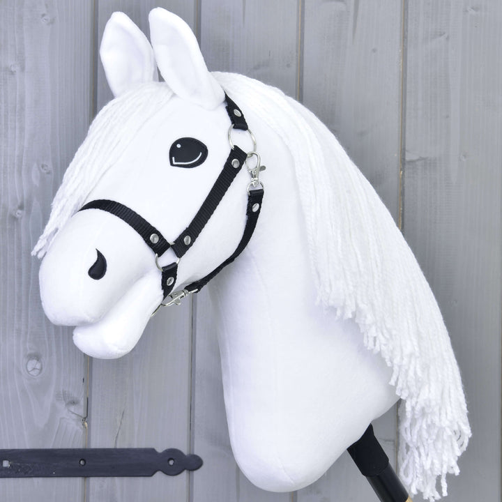 A white hobby horse with a black bridle and long white yarn mane is mounted on a stick, set against a gray wooden wall.