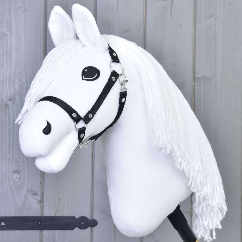 A white hobby horse with a black bridle and long white yarn mane is mounted on a stick, set against a gray wooden wall.