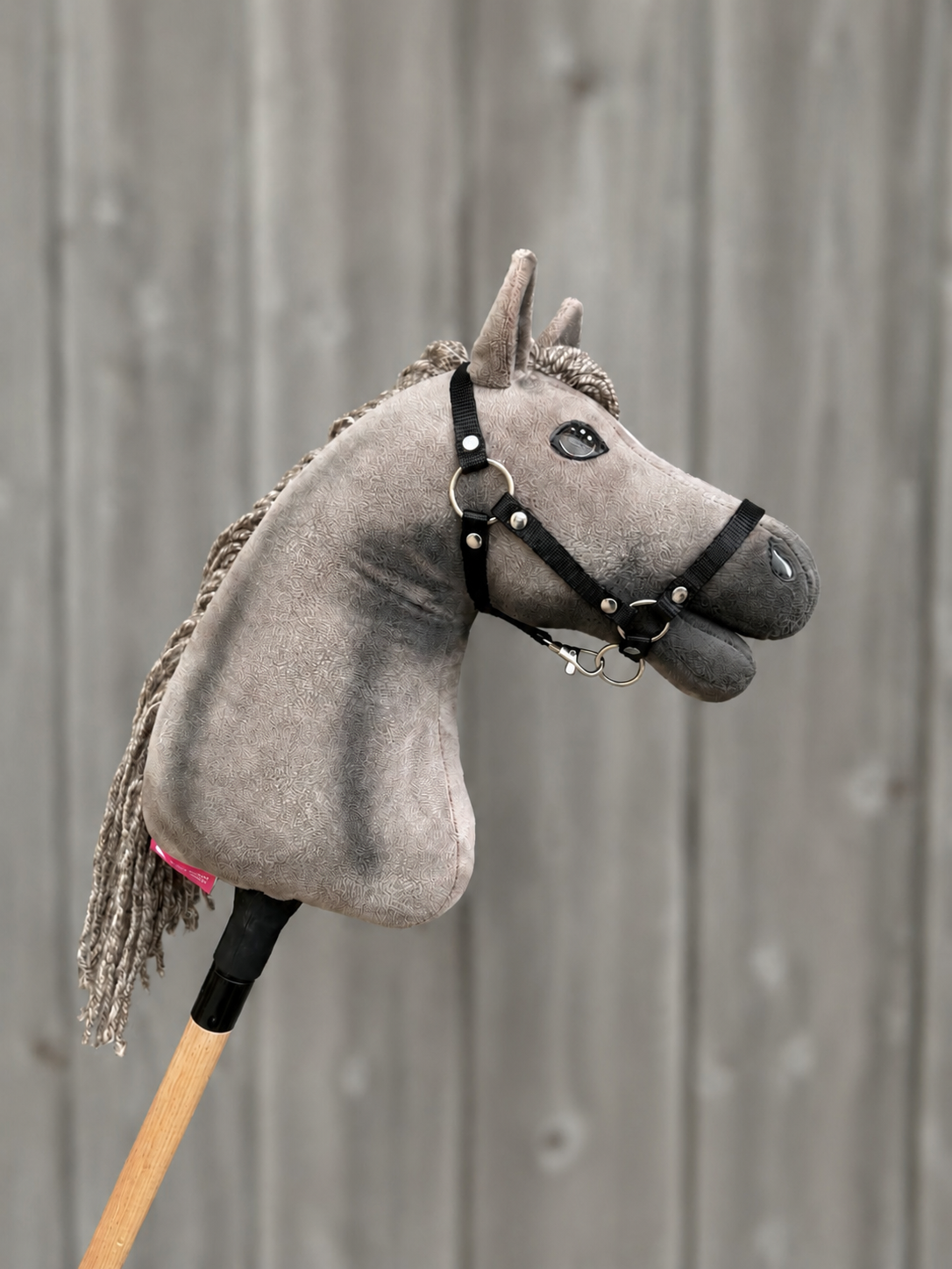 The Hobby Horse LarDen Hobby Horse Swing with Black Halter - size S features a gray Oldenburg-style stuffed head with a yarn mane and black halter on a wooden stick, shown against a blurred wooden fence background.