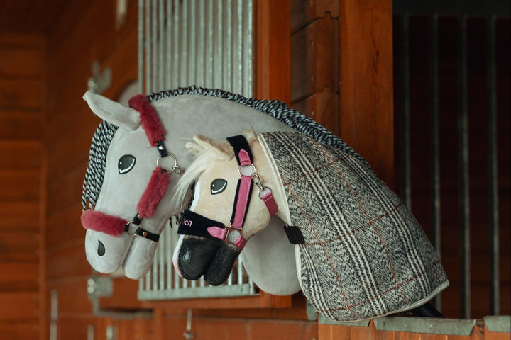 Two plush hobby horse toys with bridles and blankets rest on a wooden stable partition, resembling real horses in a stall. Perfect for hobby horsing fans, the white and gray horses have detailed features and tack.