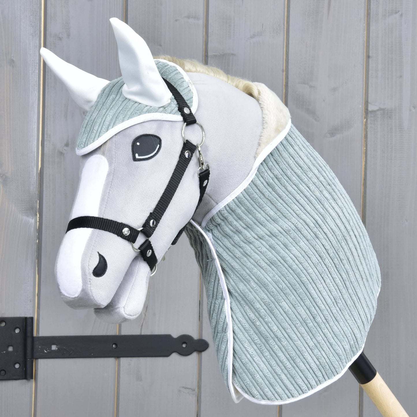 The Set Manchester Little Green by Hobby Horse LarDen features a gray and white hobby horse with a soft fabric head, black bridle, ear bonnet, and light blue-green neck cover on a wooden stick, shown against a wooden fence.