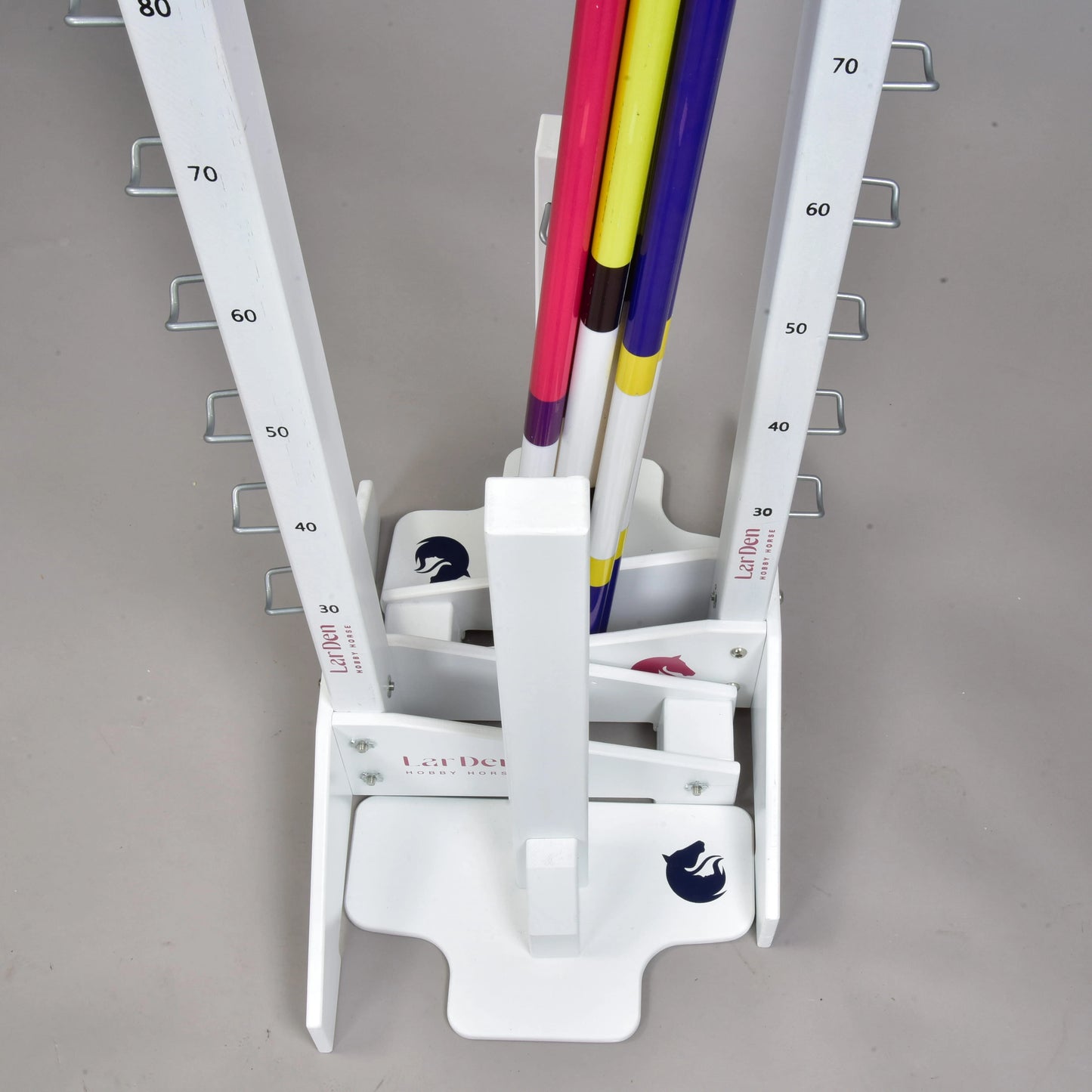 A white vertical rack with numbered markings and hooks holds colorful poles upright for adjustable jumps, ideal for hobby horsing. The base shows a horse logo and the “Hobby Horse LarDen” brand. The floor is light gray.