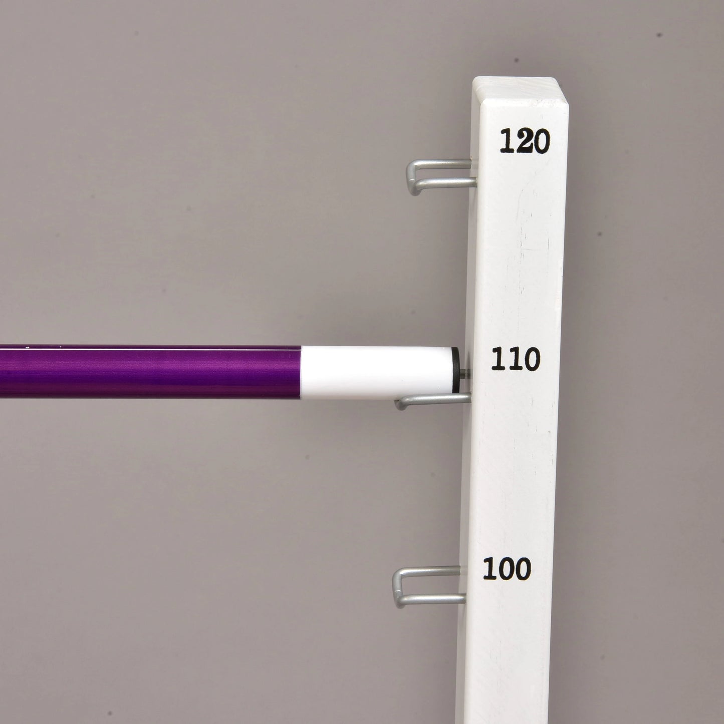 A close-up of the Hobby Horse LarDen Jump set for Hobby Horsing, showing the adjustable bar on a white stand with measurement labels (100, 110, 120), perfect for jumps—set against a plain gray background.