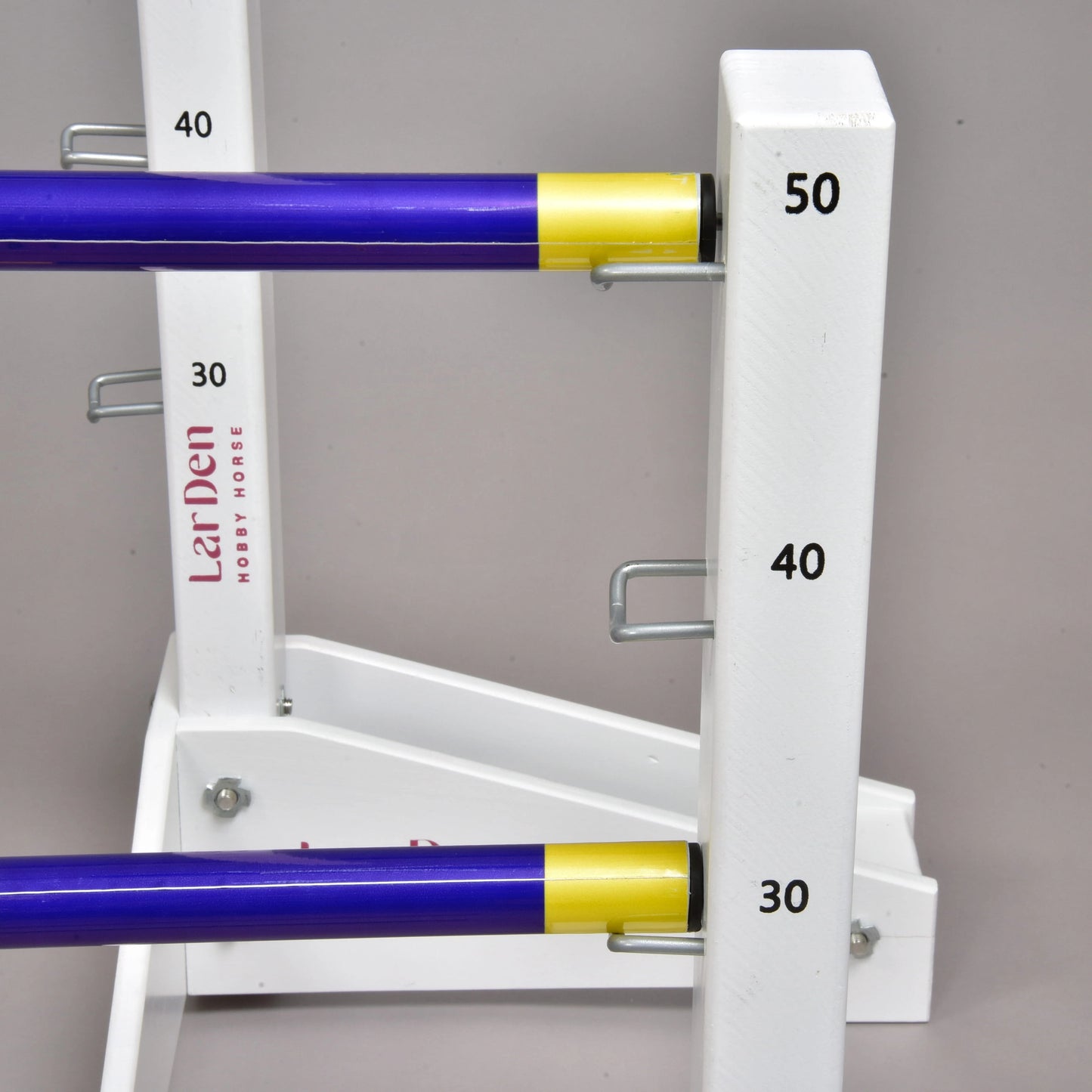 A close-up of the Hobby Horse LarDen Jump set for Hobby Horsing shows white stands with purple and yellow poles at varying heights, marked 30, 40, and 50. Metal hooks secure the adjustable poles—ideal for hobby horsing fans.