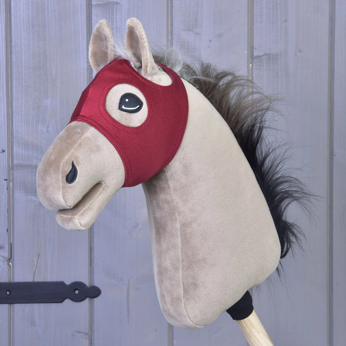 A velvet Horse Mask without ears from Hobby Horse LarDen is displayed mounted on a wooden stick in front of gray wooden paneling. Combining classic charm with practical design, this delightful, washable toy offers hours of imaginative play and easy care.