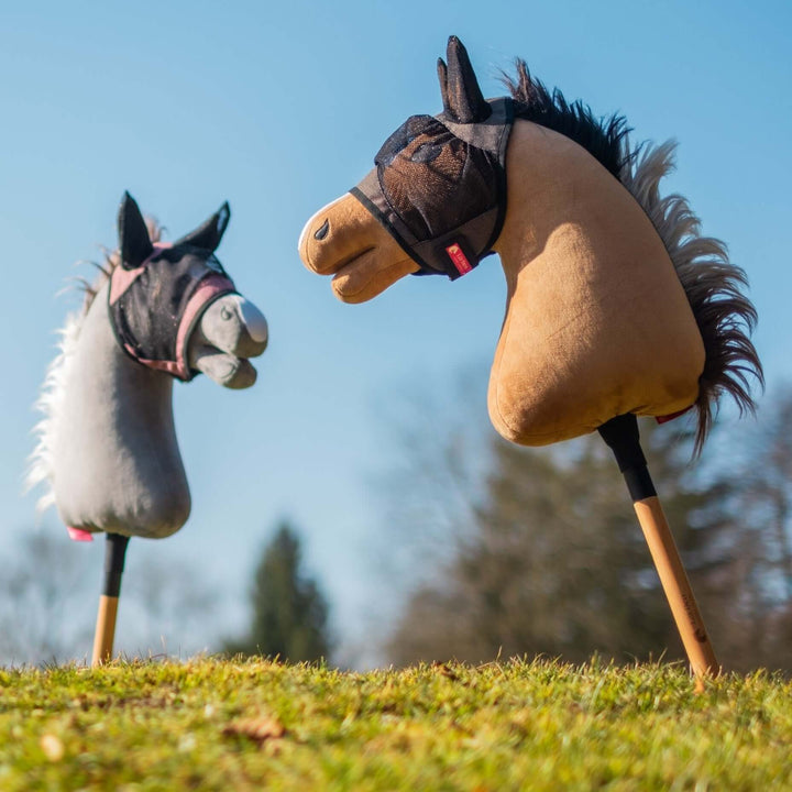 Two hobby horse toys with plush heads, one gray and one brown, stand upright on grassy ground against a clear blue sky and blurred trees. Both horses wear small fabric bridles, ready for imaginative hobby horsing adventures.