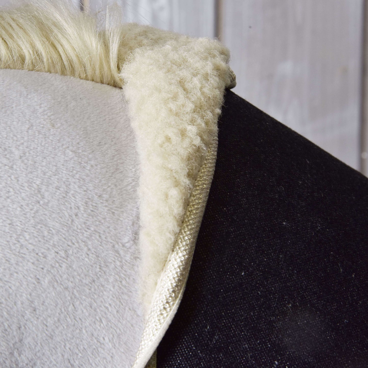 Close-up van het zadeldekje van "Set Blue Denim" van Hobby Horse LarDen met een zachte, pluizige witte schapenvachtstructuur op de rug van een donker paard, met een houten wand op de achtergrond.