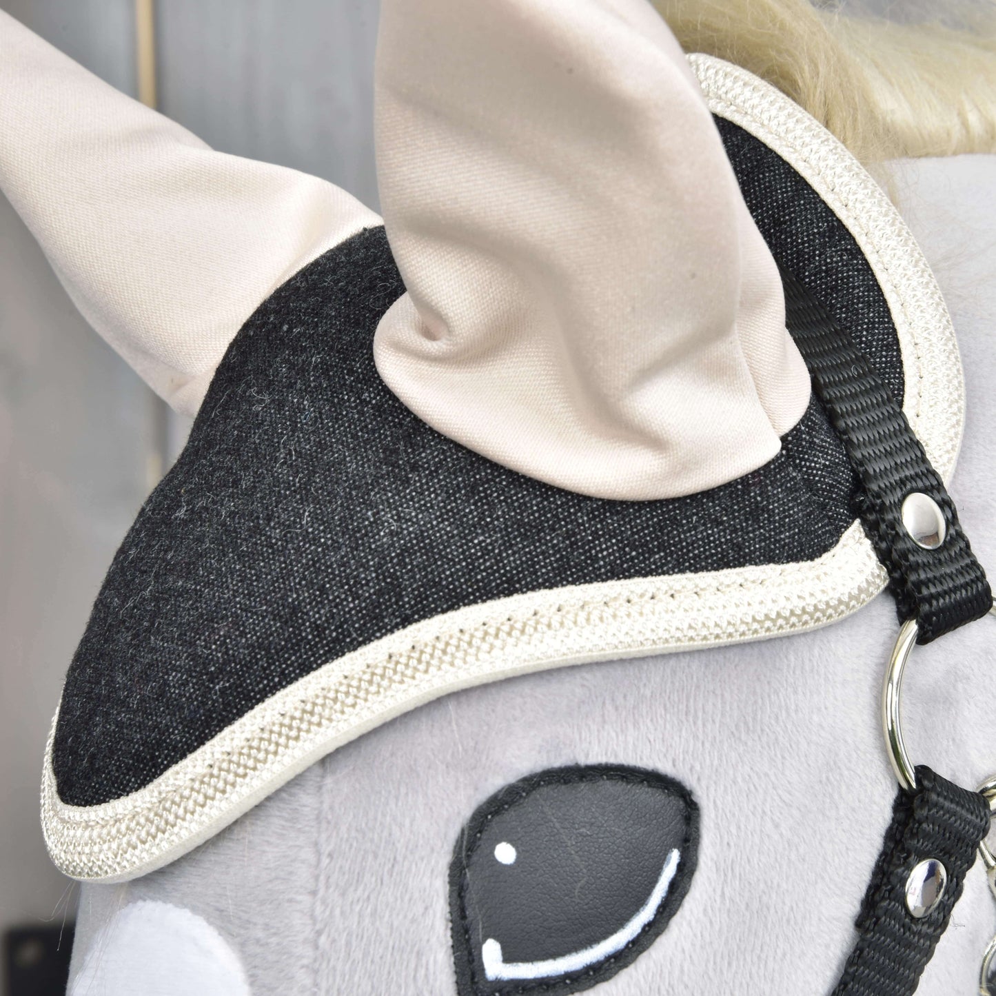 Close-up van het hoofd van Set Blue Denim van Hobby Horse LarDen met een zwart hoofdstel met witte rand, crèmekleurige oren, een glad grijs oppervlak en een geborduurd oog, als decoratie voor paardenliefhebbers.