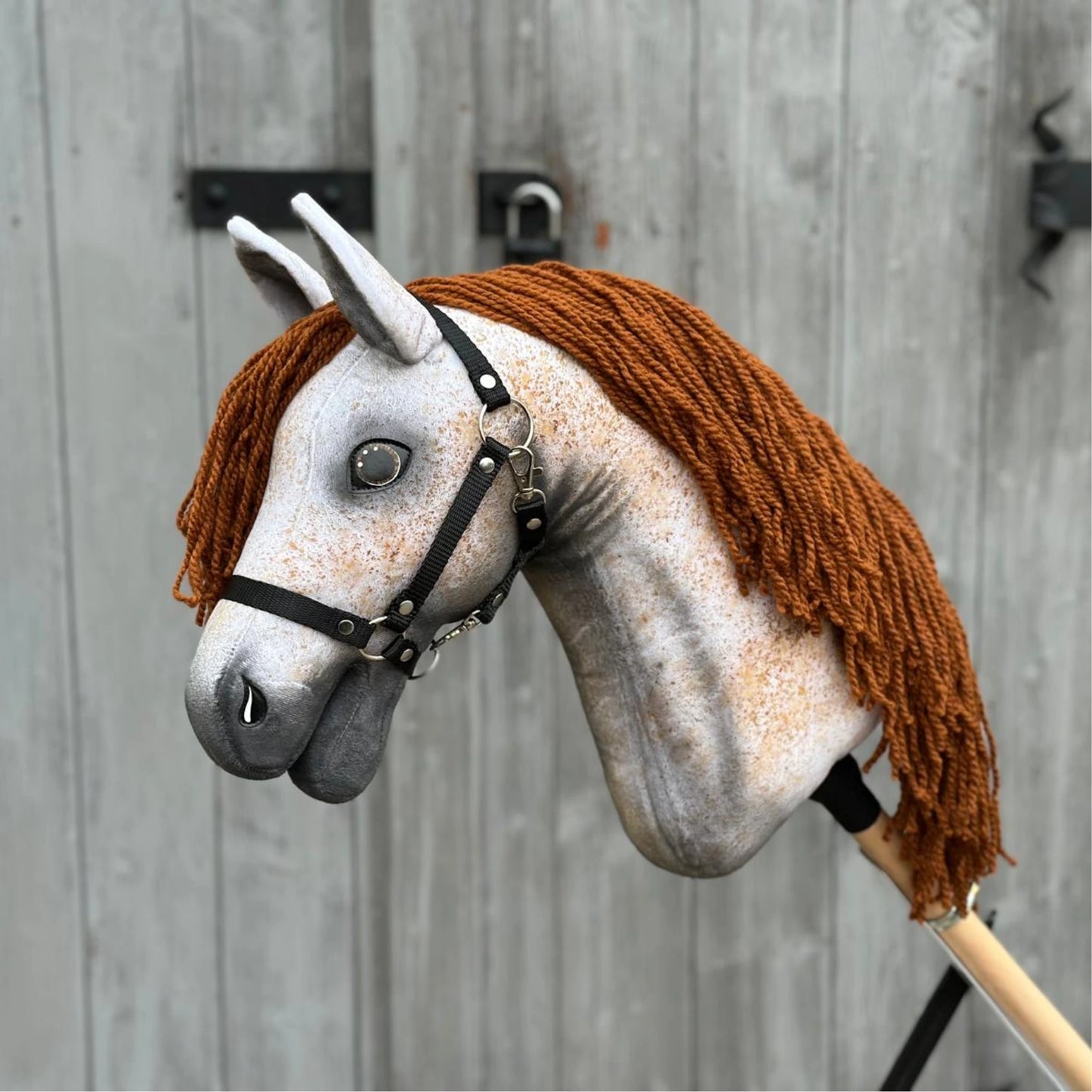 Hobby Horse Pompo met zwart halster van Hobby Horse LarDen met een grijs gespikkeld gezicht, bruine manen van garen en een zwart hoofdstel, geïnspireerd op de Hannoveraanse hengst, getoond tegen een verweerd houten hek.