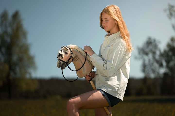 A young person with long blond hair, wearing a light jacket and shorts, enjoys hobby horsing by riding a hobby horse outdoors in a sunny, grassy field with trees in the background.
