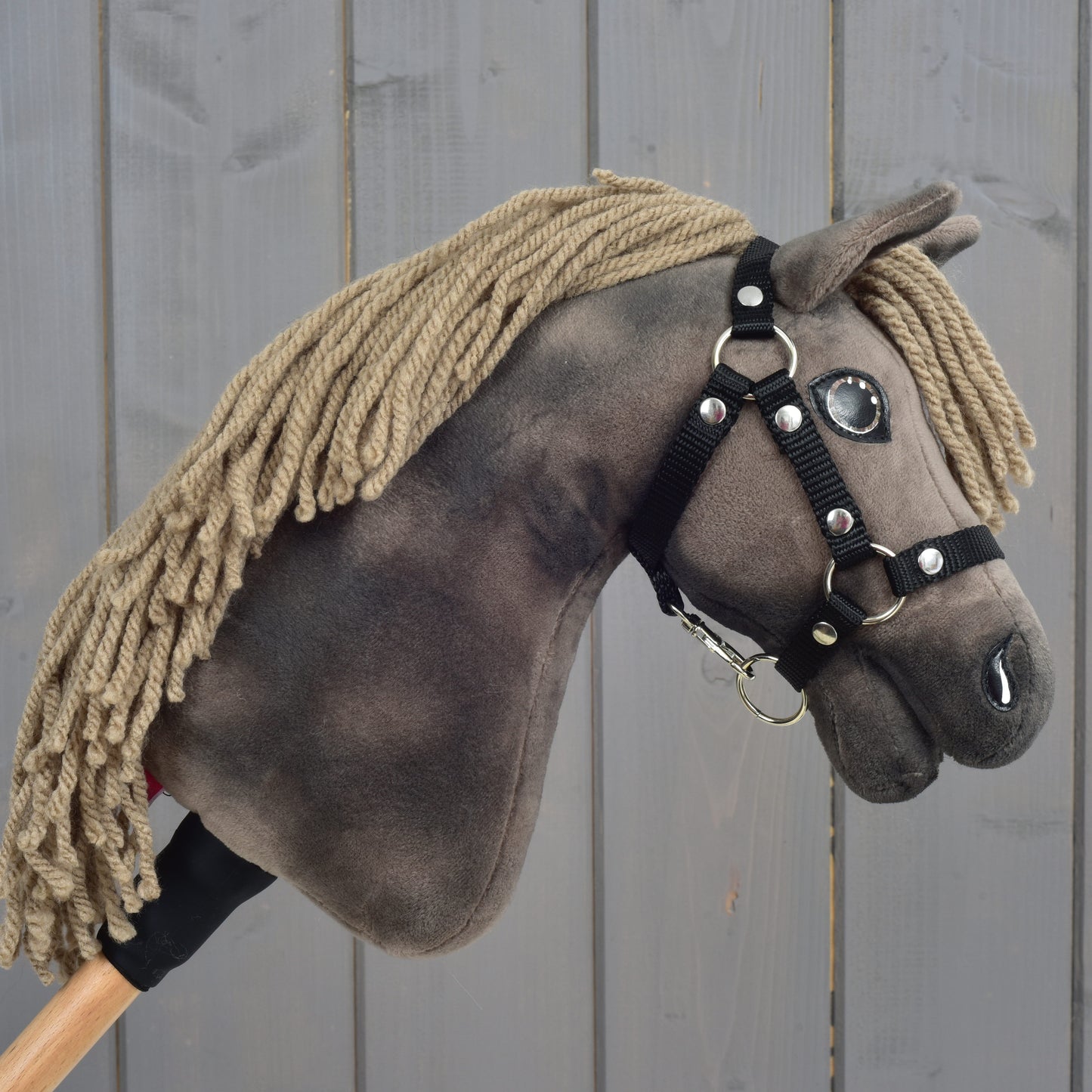 Close-up van Hobby Horse Avalon met zwart halster, maat XS van Hobby Horse LarDen, met een pluchen sportpaardontwerp, bruine manen van garen, knoopogen en een zwart hoofdstel op een houten stok, tegen een grijze houten achtergrond.