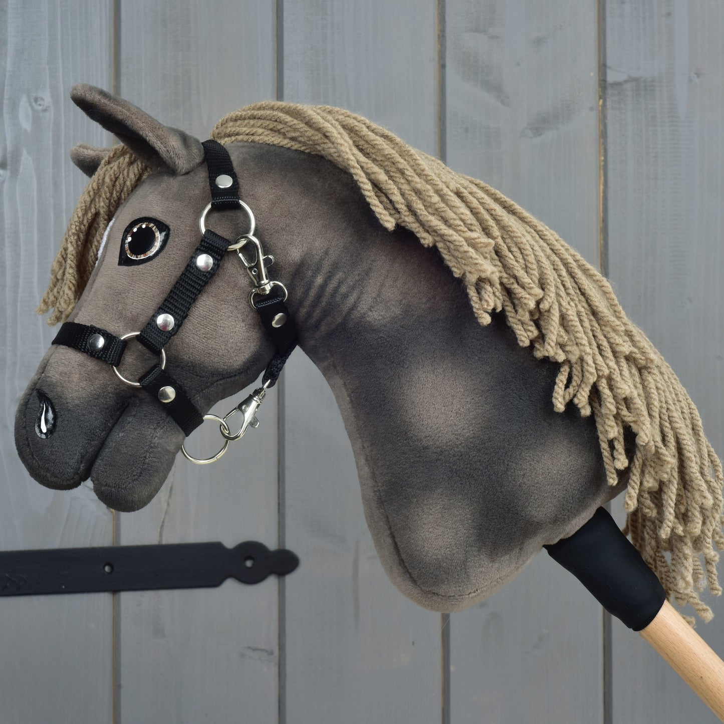 Hobby Horse Avalon met zwart halster, maat XS van Hobby Horse LarDen, een handgemaakte Hobby Horse geïnspireerd op een Engelse Warmbloed sportpaard. Met lichtbruine manen van garen, een donkergrijze stoffen kop en bevestigd op een houten stok met een strak zwart halster. Gefotografeerd tegen een grijze houten wand, ideaal als fantasierijk springspeelgoed.