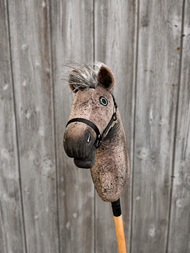 Hobby Horse Bolero met zwart halster - maat S van Hobby Horse LarDen, met een bruine pluchen kop, grijze manen en een houten stok, getoond voor een verweerd hek.