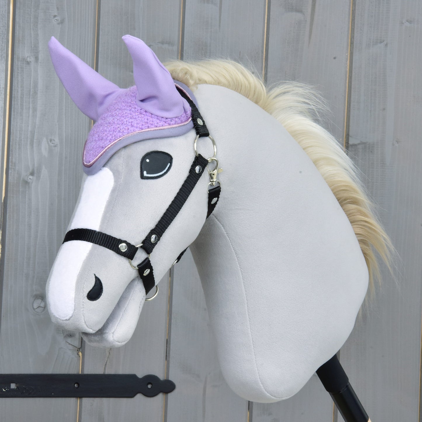 A plush hobby horse with a light gray body, black bridle, and cream mane wears the Hobby Horse LarDen Ear bonnet Orchid for insect protection. The background shows a wooden fence.