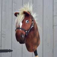 Hobby Horse Cinnamon met zwart halster - maat S van Hobby Horse LarDen, een bruin pluchen Hobby Horse met een witte bles, zwart halster en pluizige blonde manen, bevestigd op een houten stok voor een grijs houten hek.