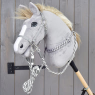 Een grijze pluchen Hobby Horse van Hobby Horse LarDen met een witte bles, uitgerust met Set Start zilver van Hobby Horse LarDen met halster voor Hobby Horse, cordeo, leadrope en teugels, is bevestigd op een houten stok en leunt tegen een grijze houten muur.