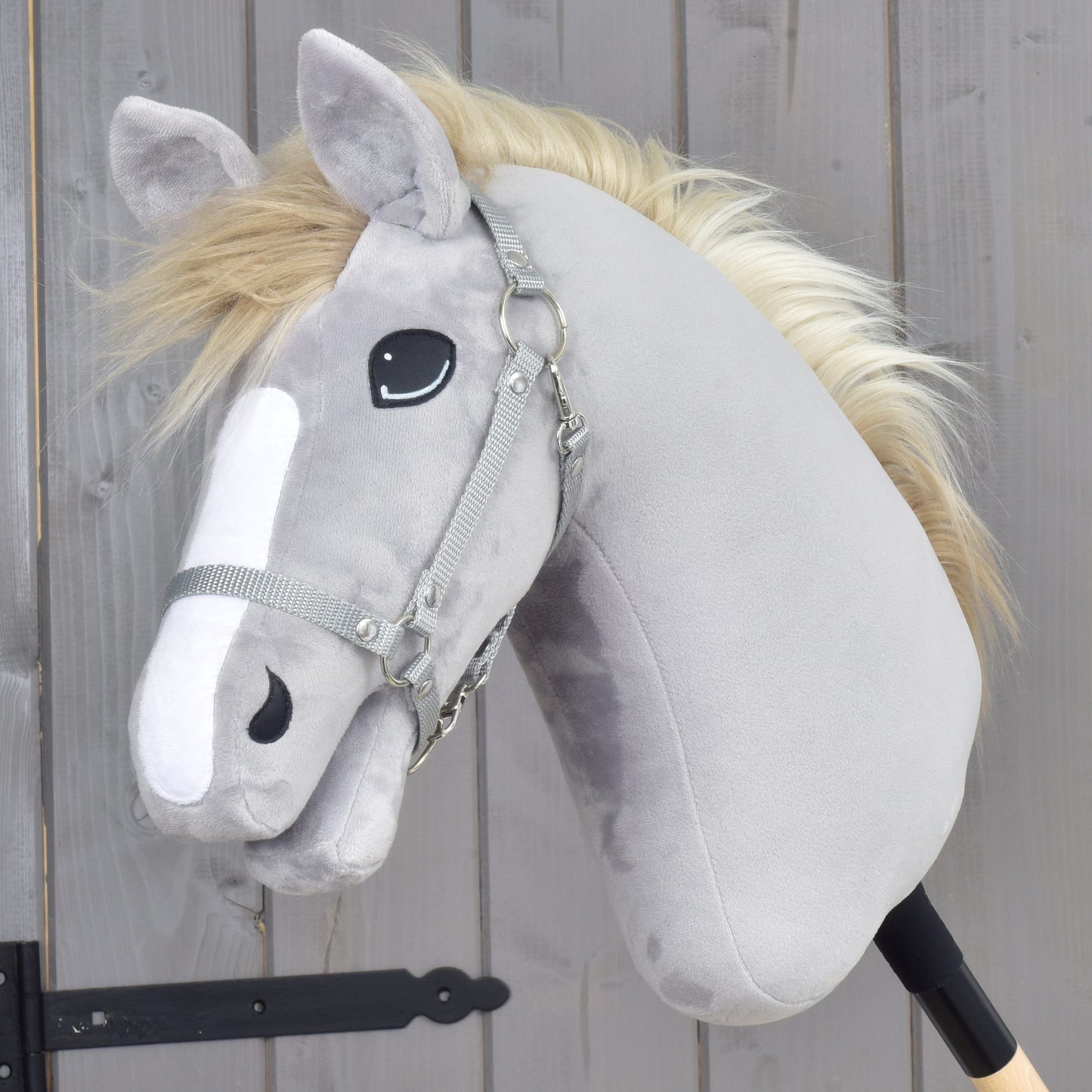 A grey plush hobby horse with a white mane and bridle is mounted on a stick in front of a wooden fence, featuring realistic reins and the stylish Set Start Silver (halter + cordeo + lead rope + reins) from Hobby Horse LarDen.
