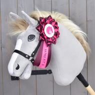 A plush Hobby Horse Ribbon Raspberry Rose by Hobby Horse LarDen, featuring a gray face, cream mane, and black bridle mounted on a stick, is adorned with a large pink and white dressage ribbon displaying the LarDen logo on its neck. The background consists of wooden paneling.