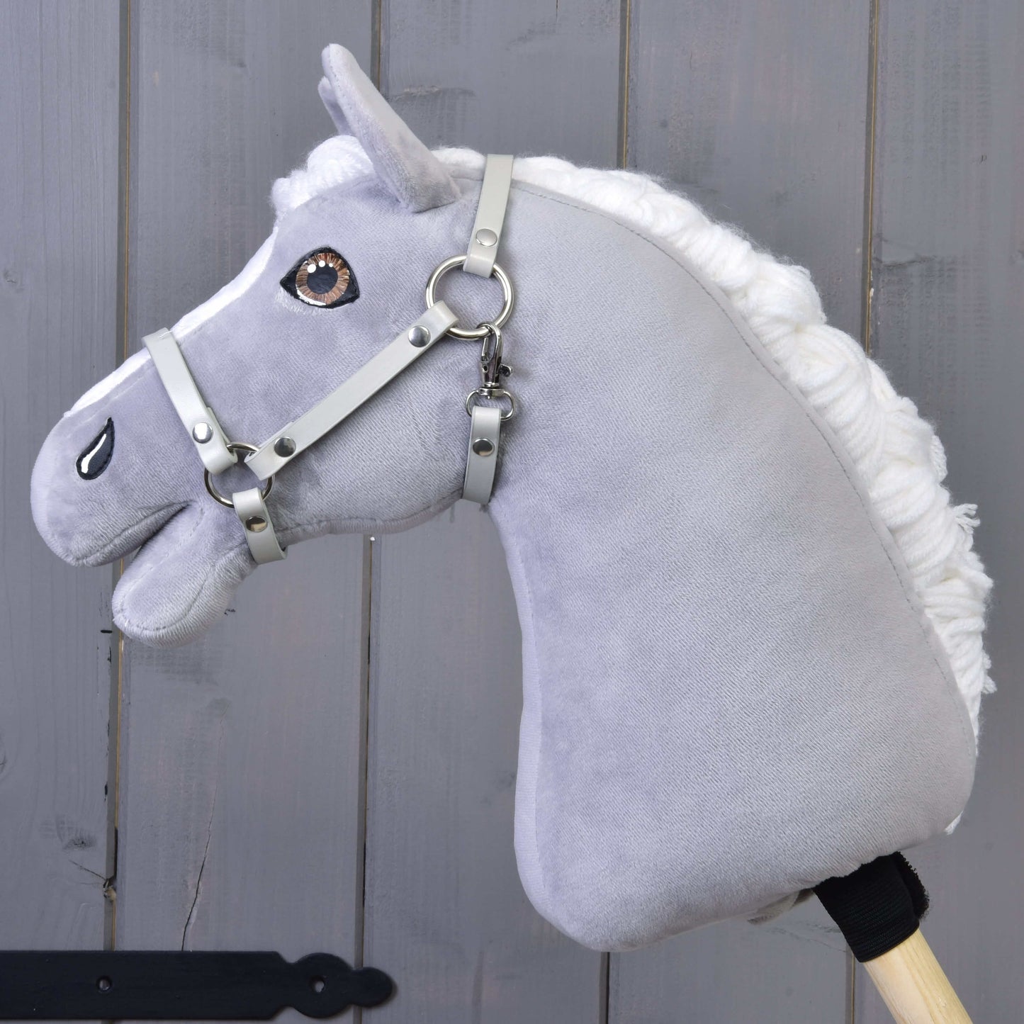 The Hobby Horse LarDen features a gray plush horse with a white mane, brown eyes, and a Halter in light grey leatherette (size S), mounted on a wooden stick and shown against a gray wooden background.
