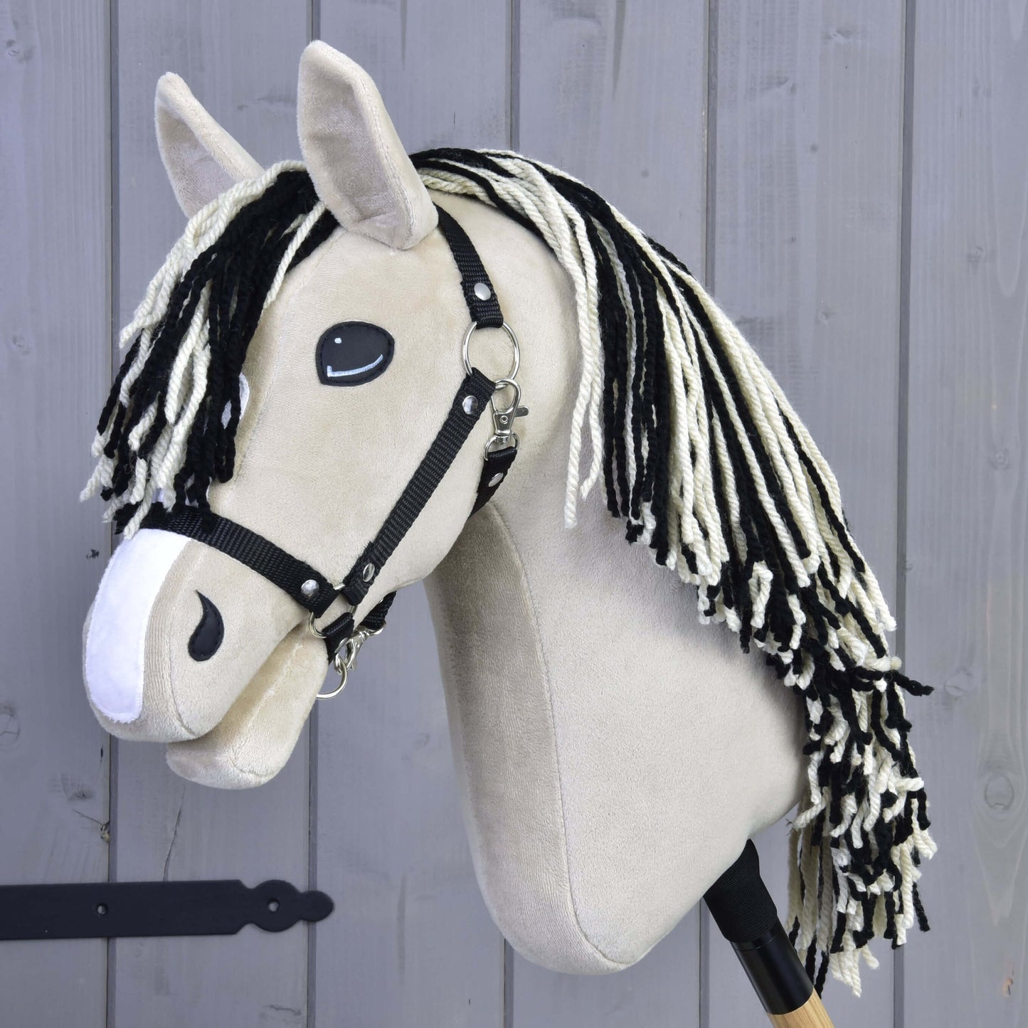 Hobby Horse Aurora met zwart halster van Hobby Horse LarDen, geïnspireerd op een valkkleurige merrie, met zwart-witte manen van garen en zwart halster, afgebeeld voor een grijze houten wand.