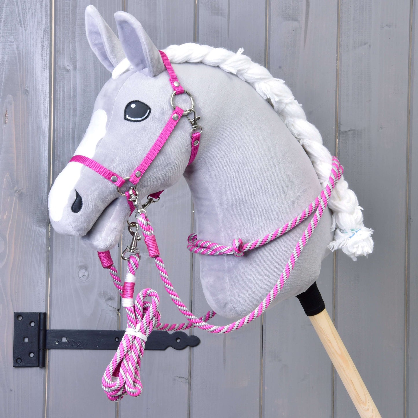 Set Start roze (halster + cordeo + lead rope + teugels) van Hobby Horse LarDen op een grijze pluchen Hobby Horse met witte manen, gemonteerd op een houten stok tegen een grijze houten wand.