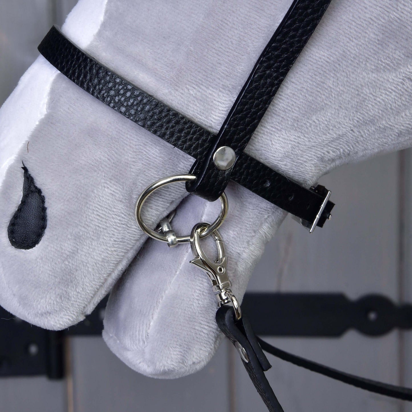 Close-up van een witte pluchen paardenkop met het Compleet zwart hoofdstel met diamanten van Hobby Horse LarDen, met afneembare teugels en een Engelse neusriem met metalen bit, tegen een houten achtergrond met zichtbaar donker beslag.