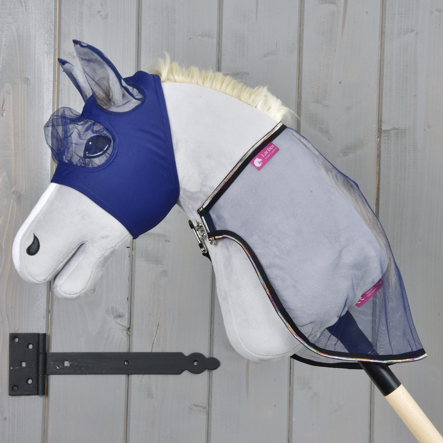 Set Vlieg Combo van Hobby Horse LarDen met een pluchen Hobby Horse met een blauw vliegmasker en mesh vliegendeken op een houten stok, tegen een grijze houten wand, klaar voor je volgende avontuur.