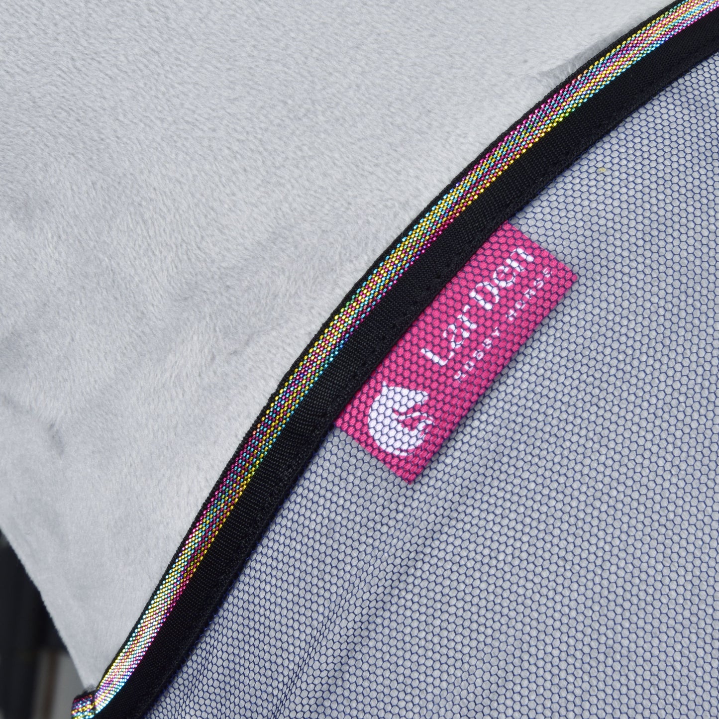 Close-up van meshstof met een roze Set Vlieg Combo-label met kattengezicht-icoon van Hobby Horse LarDen, afgewerkt met veelkleurige rand, een stijlvol accessoire voor je Hobby Horse, weergegeven op een lichtgrijze achtergrond.