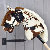 Hobby Horse Dakota met zwart halster van Hobby Horse LarDen met bruine, zwarte en witte vlekken, een bruine manen van garen en een zwart halster. Gemonteerd op een houten stok tegen een grijze muur, geschikt voor elk geslacht.