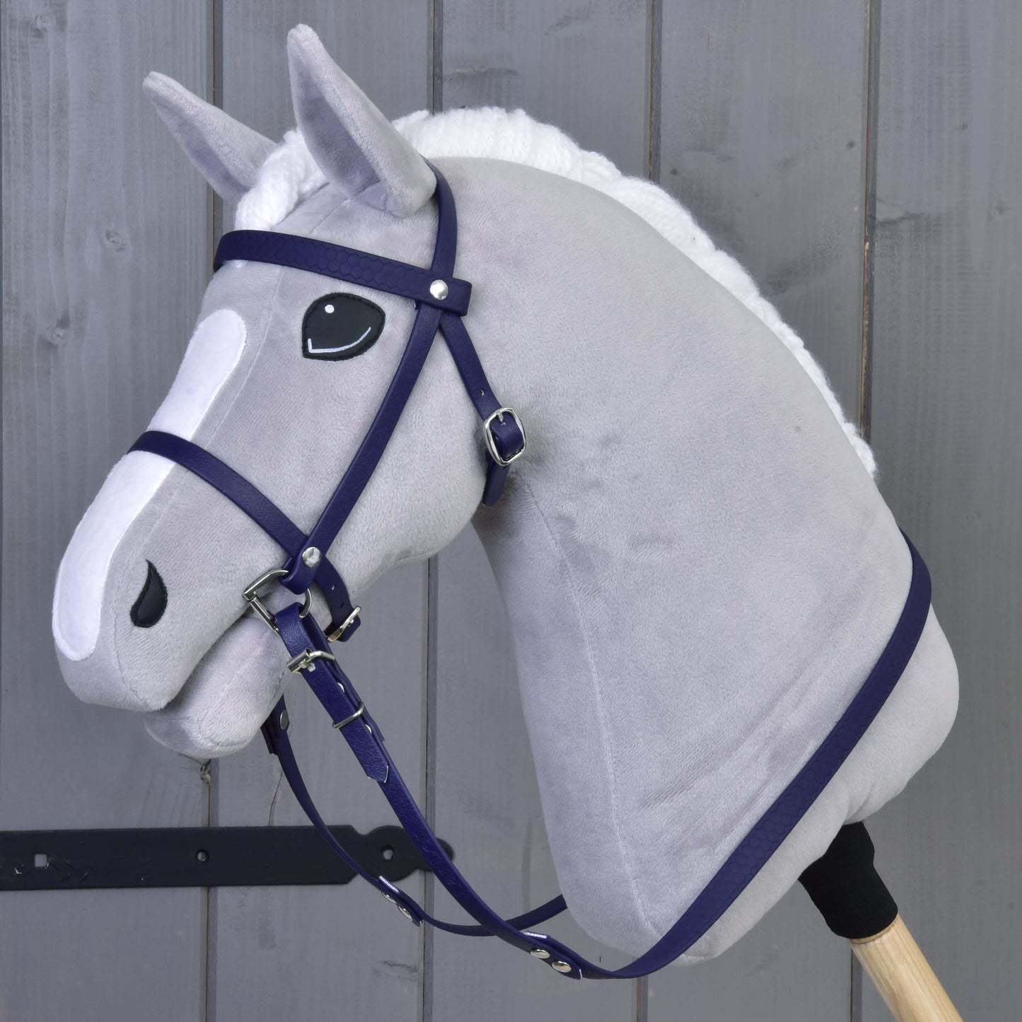 Een grijze pluchen Hobby Horse van Hobby Horse LarDen, met een compleet hoofdstel met neusriem in donkerpaars, zwart-witte ogen, witte manen en gemonteerd op een houten stok, staat voor een grijze houten wand.