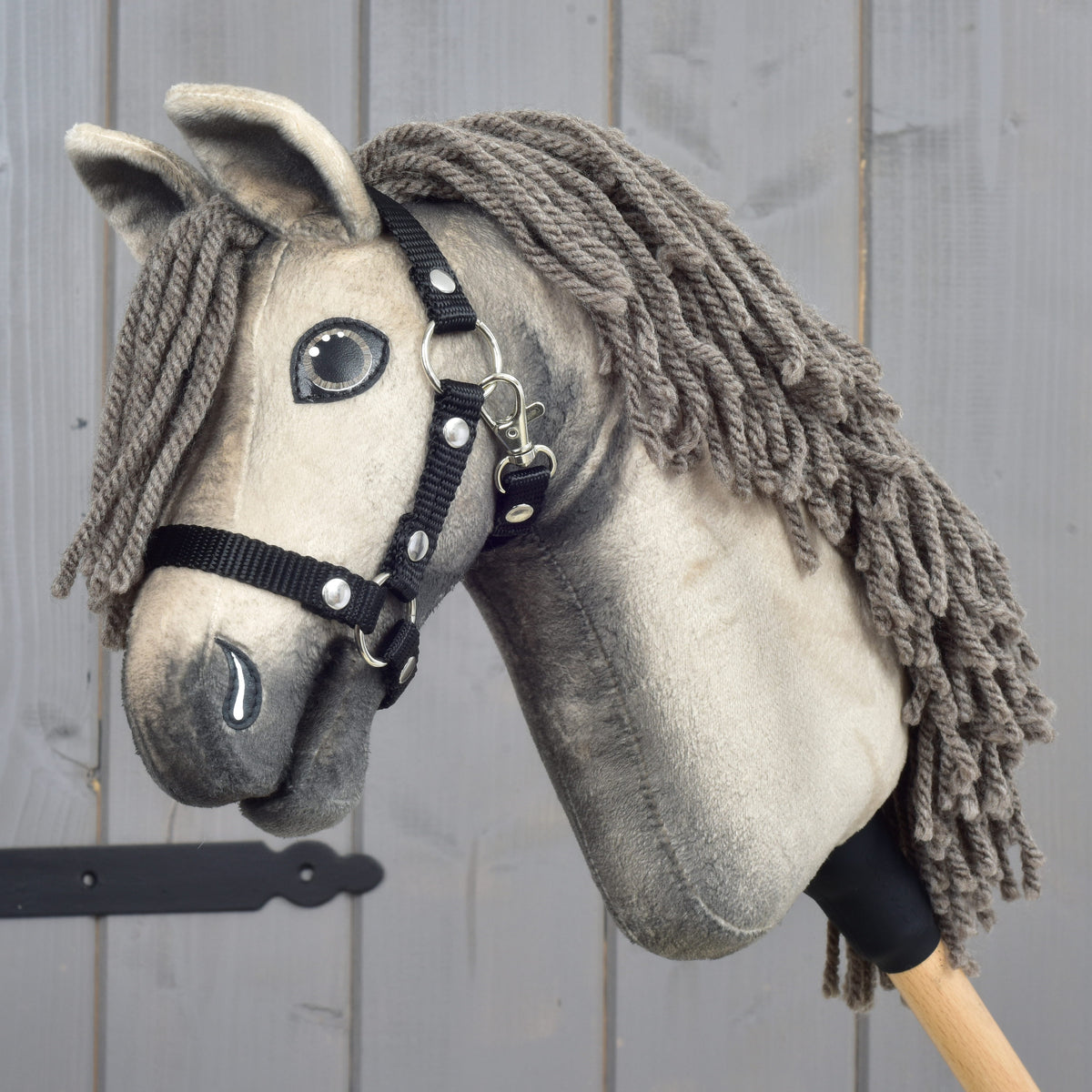 The Hobby Horse LarDen Hobby Horse Dusty with Black Halter (size XS), featuring a plush Hanoverian design with a gray yarn mane, is displayed in front of a gray wooden background.