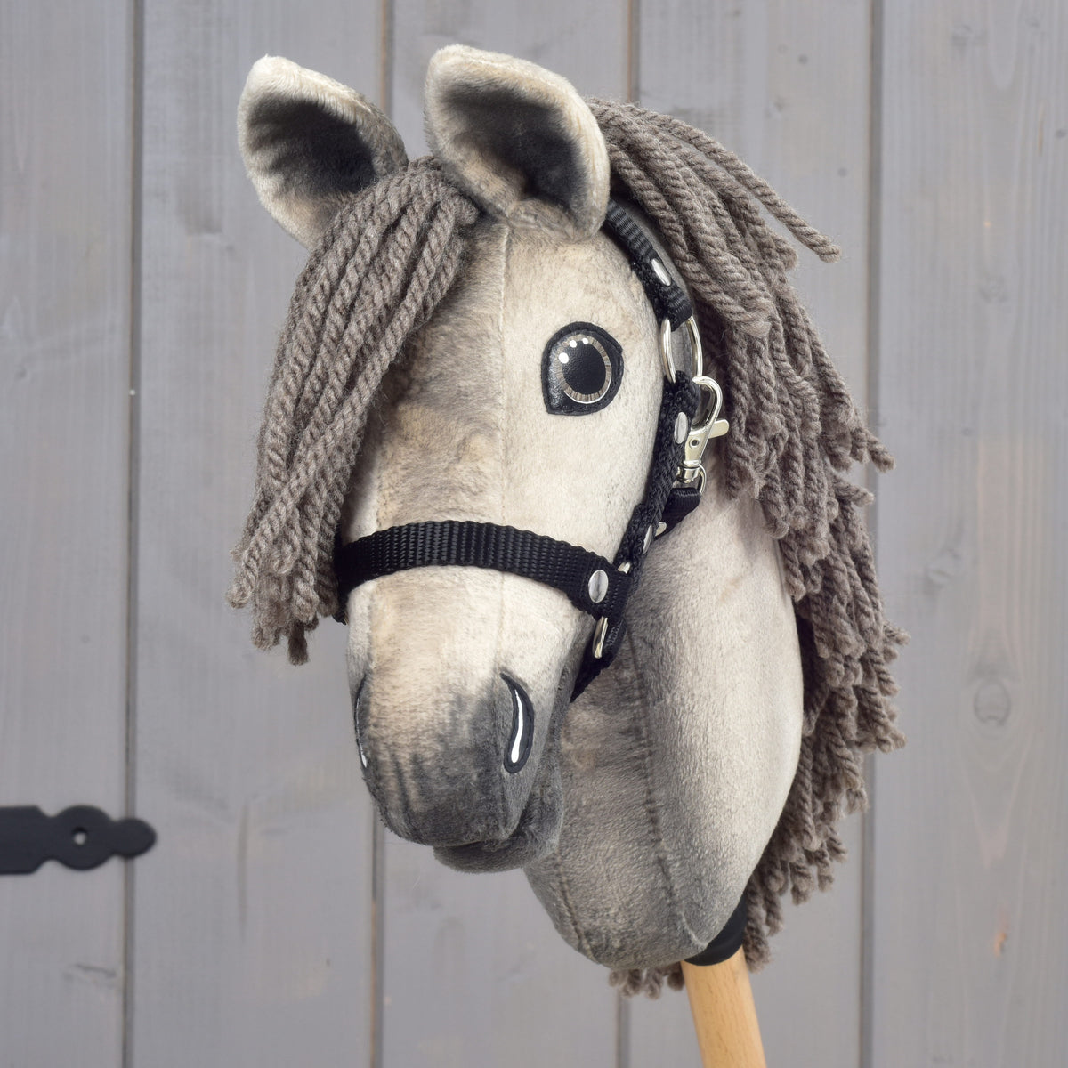 A close-up of the Hobby Horse LarDen "Hobby Horse Dusty with Black Halter" in size XS, modeled after a Hanoverian stallion, featuring a gray yarn mane, black halter, and expressive button eyes, mounted on a wooden stick against a gray wooden background.