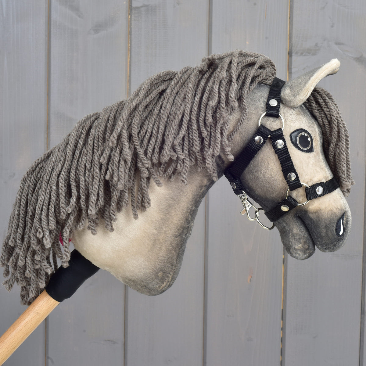 A Hobby Horse LarDen "Hobby Horse Dusty with Black Halter" in size XS, featuring a gray yarn mane and a wooden stick handle, is displayed in front of a gray wooden fence.