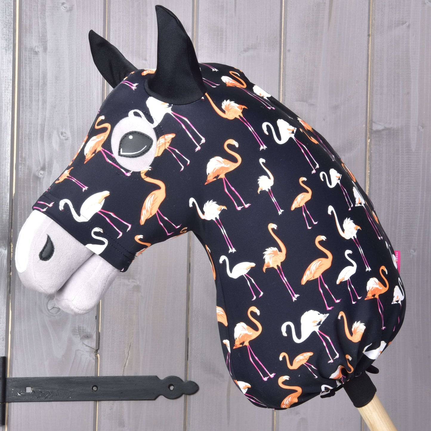 Beschermpak Lycra Flamingo van Hobby Horse LarDen heeft een lycra hoes met oranje en witte flamingo's op een donkere achtergrond, gemonteerd op een houten stok tegen een wand met houten panelen.