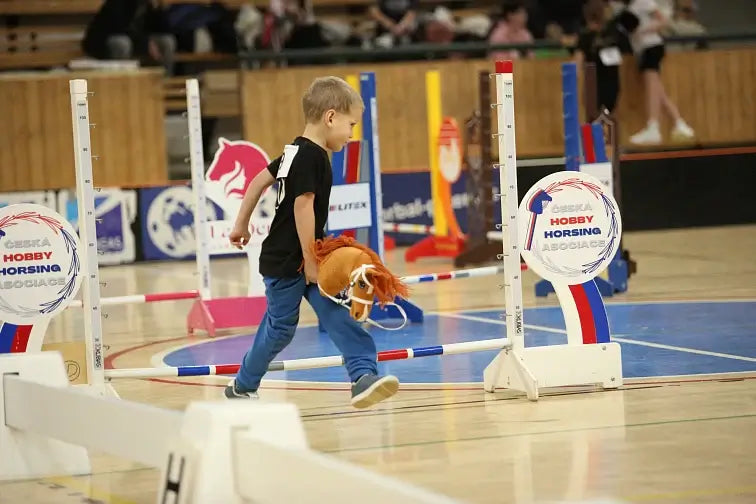 Een jonge jongen neemt deel aan een Hobby Horse-wedstrijd en loopt met een speelgoedpaard over een gymvloer, omringd door kleurrijke springhindernissen en borden van een Hobby Horsing-evenement.