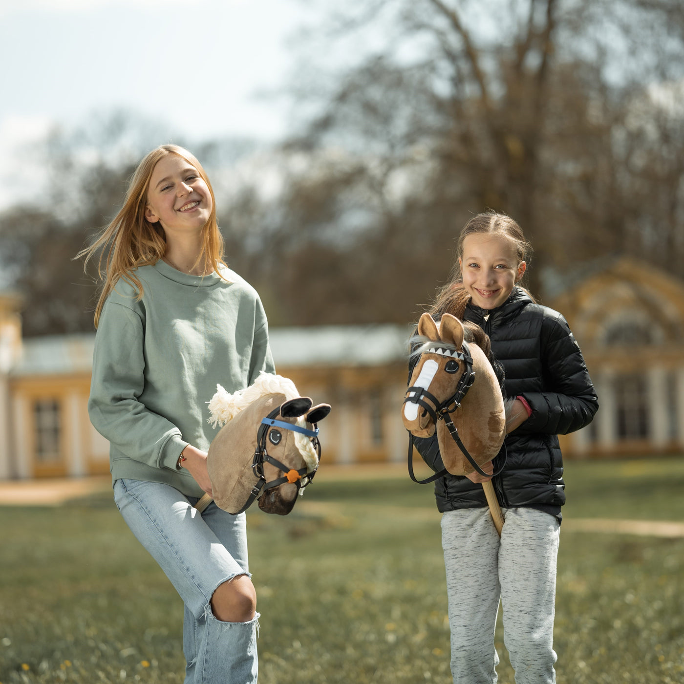 Twee glimlachende meisjes staan buiten met hun hobby horses in de hand. De een draagt een lichtgroene trui en gescheurde jeans, de ander een zwart jack en grijze broek. Hobby Horsing brengt hun plezier terwijl ze poseren bij een groot gebouw met bomen op de achtergrond.