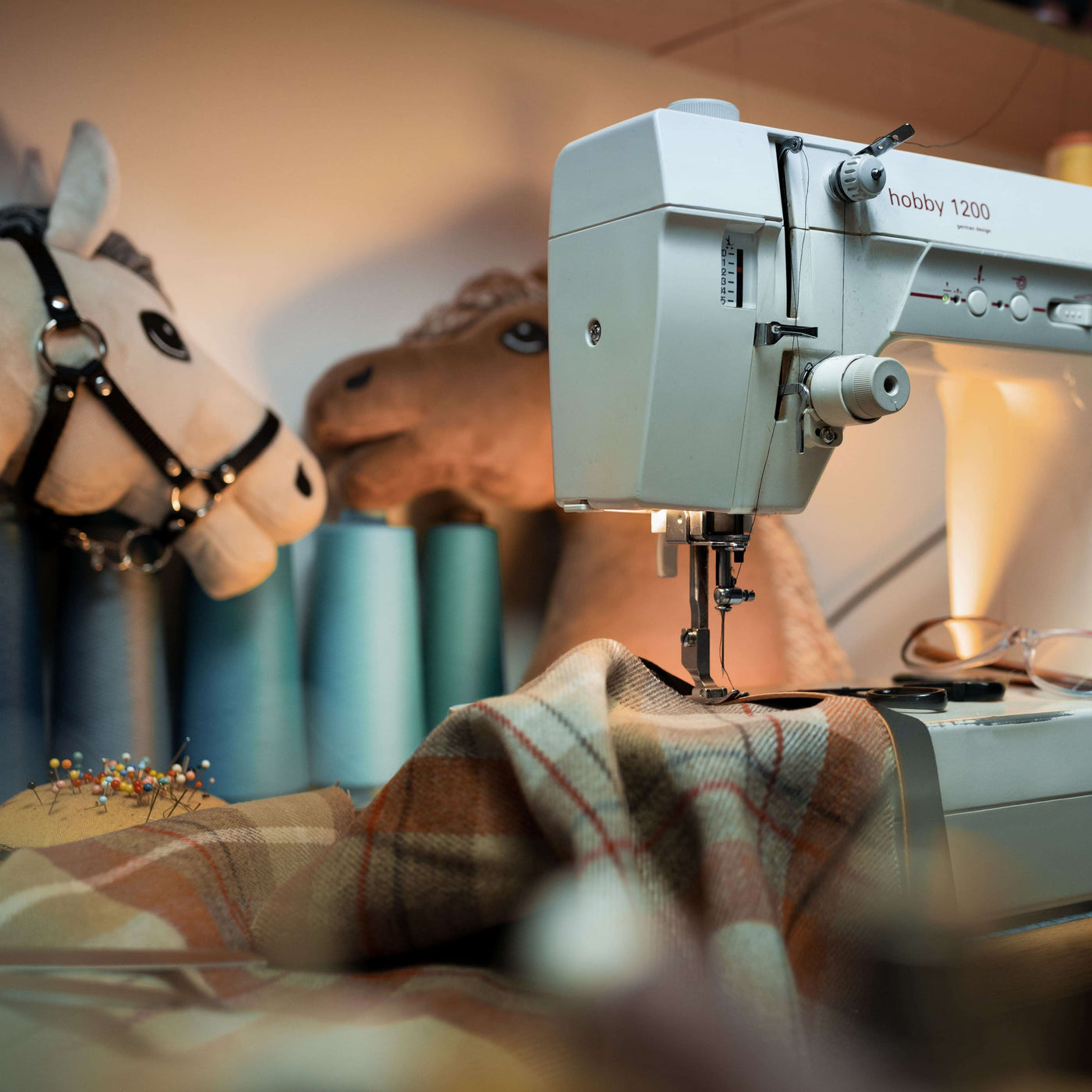 A sewing machine stitching plaid fabric, with large spools of blue thread and two plush hobby horse toys in the background, creating a cozy crafting atmosphere.