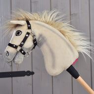 Hobby Horse Ivory Dream met zwart halster van Hobby Horse LarDen, maat XS, met zachte ivoorkleurige pluche en een houten stok, leunt tegen een grijze houten wand.