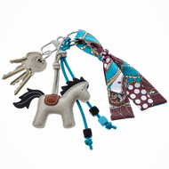 Een set sleutels aan een ring met de Hobby Horse LarDen Sleutelhanger Horse Grey, blauwe koordjes met kralen en een kleurrijke blauw-, bruin- en witdessin sjaal die aan de sleutelhanger is geknoopt.