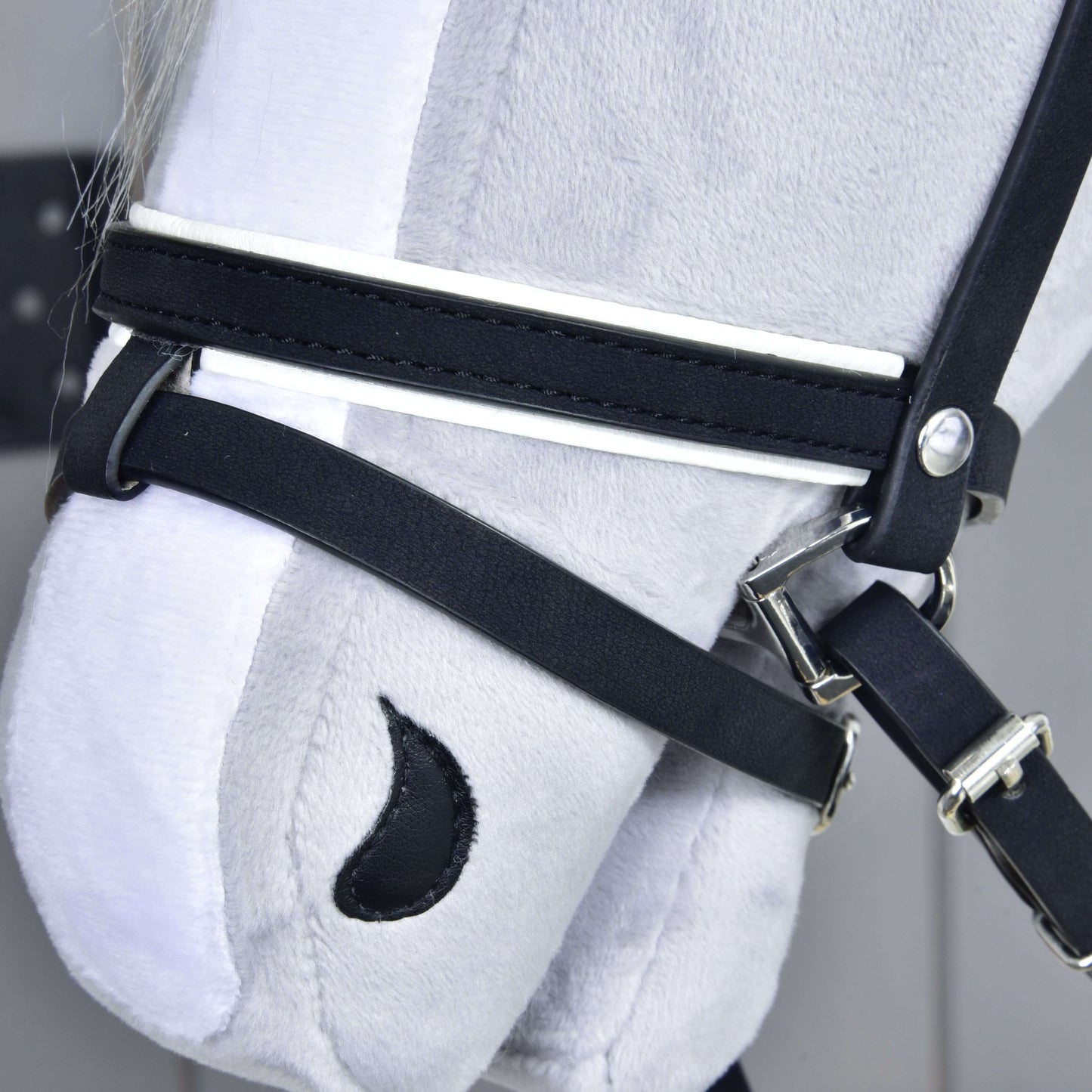 Close-up van een grijze pluchen paardenkop van Hobby Horse LarDen met het Complete De Luxe hoofdstel met 2 frontriemen. Geborduurde details zoals een zwart halvemaanvormig neusgat vallen op tegen een lichte achtergrond.