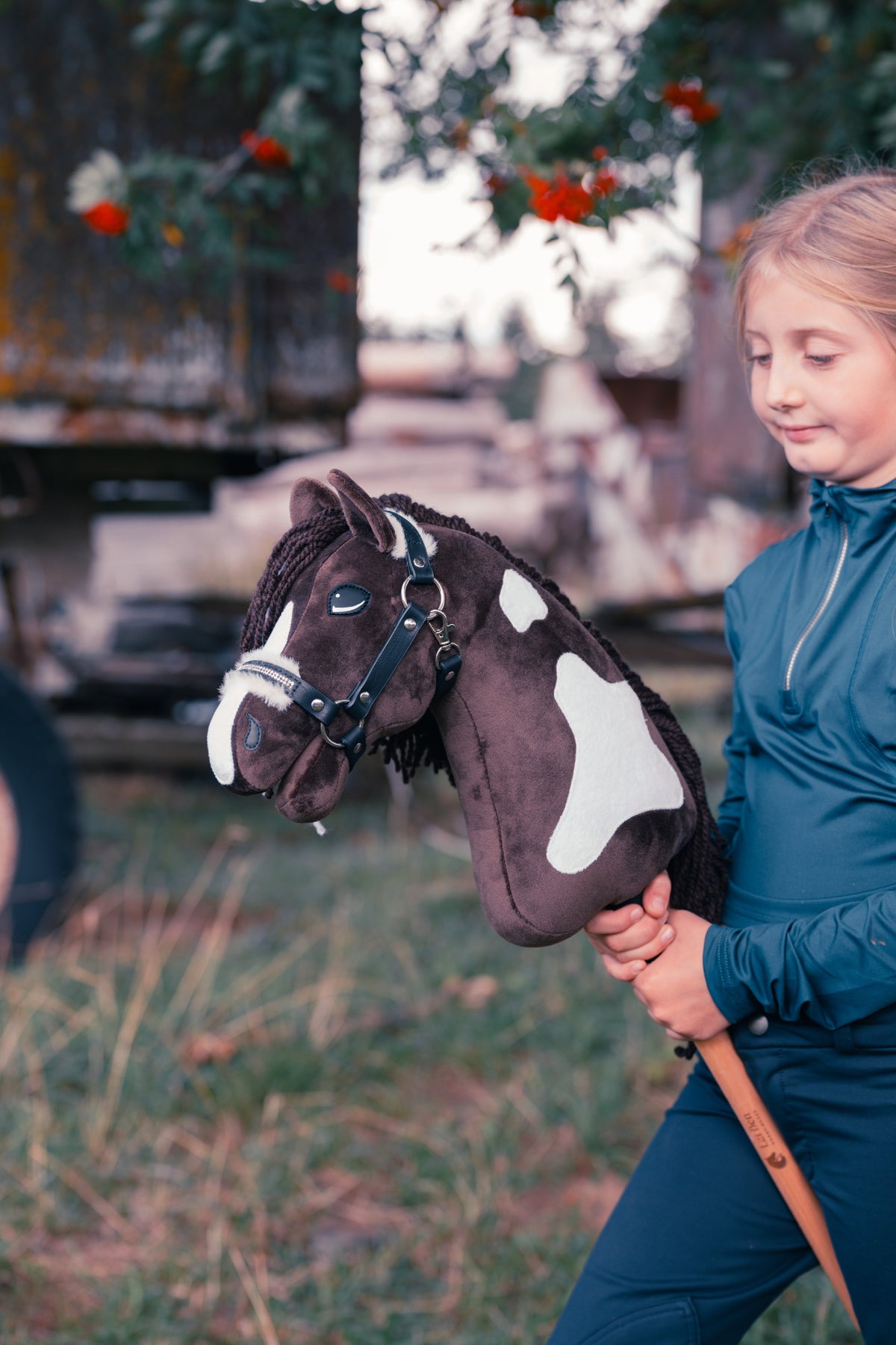 Jonge deelnemer in donkerblauwe rijkleding houdt Hobby Horse Monte van Hobby Horse LarDen vast in een weide voor een oude houten wagen en een lijsterbes met rode bessen, met een bruin-witte vacht geïnspireerd op de Paint Horse, lange donkere manen van garen en het met bont gevoerde Halster Espresso