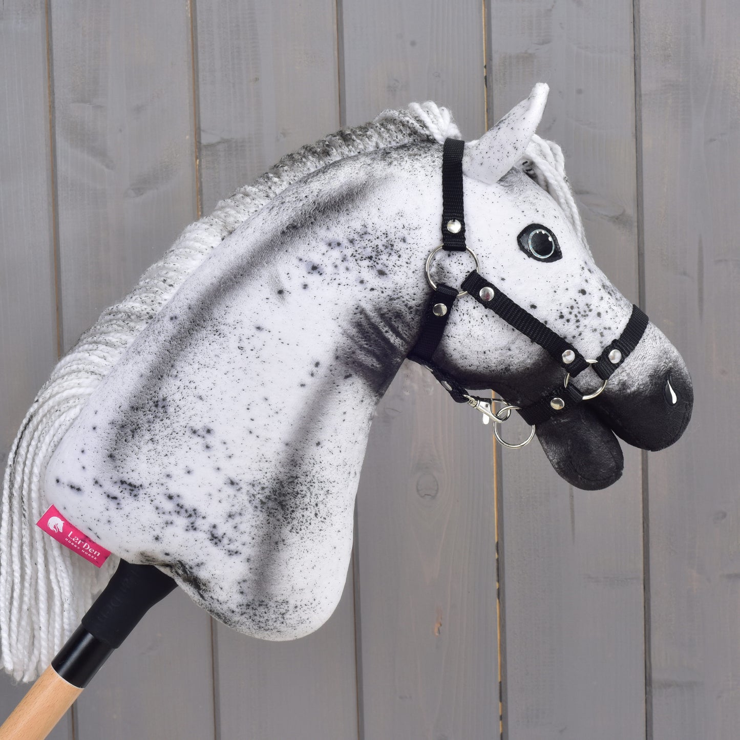 Hobby Horse Naivy met zwart halster - maat S van Hobby Horse LarDen, met een opvallend wit en grijs gespikkeld ontwerp, gemonteerd op een houten stok voor een grijze houten wand.