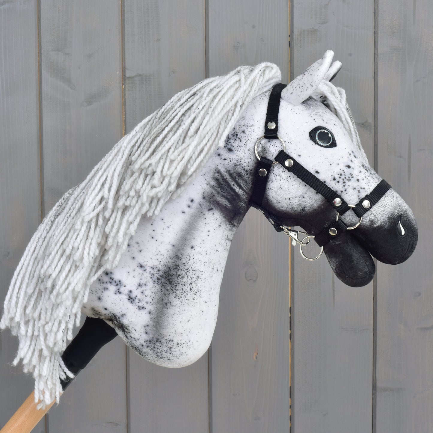 Hobby Horse Naivy met zwart halster - maat S van Hobby Horse LarDen, een handgemaakte Appaloosa Hobby Horse met witte en grijze manen van garen, opvallende zwarte vlekken en een strak zwart halster, tegen een rustieke houten achtergrond. Ideaal voor fantasierijke avonturen als hengst.